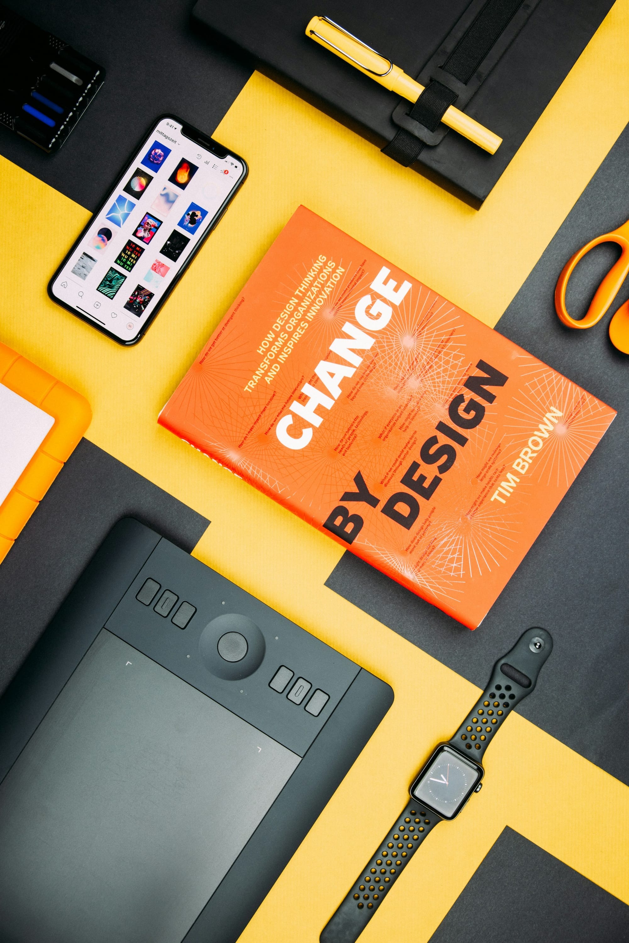A design book, smartphone, drawing tablet and stationery arranged on a yellow and black surface.