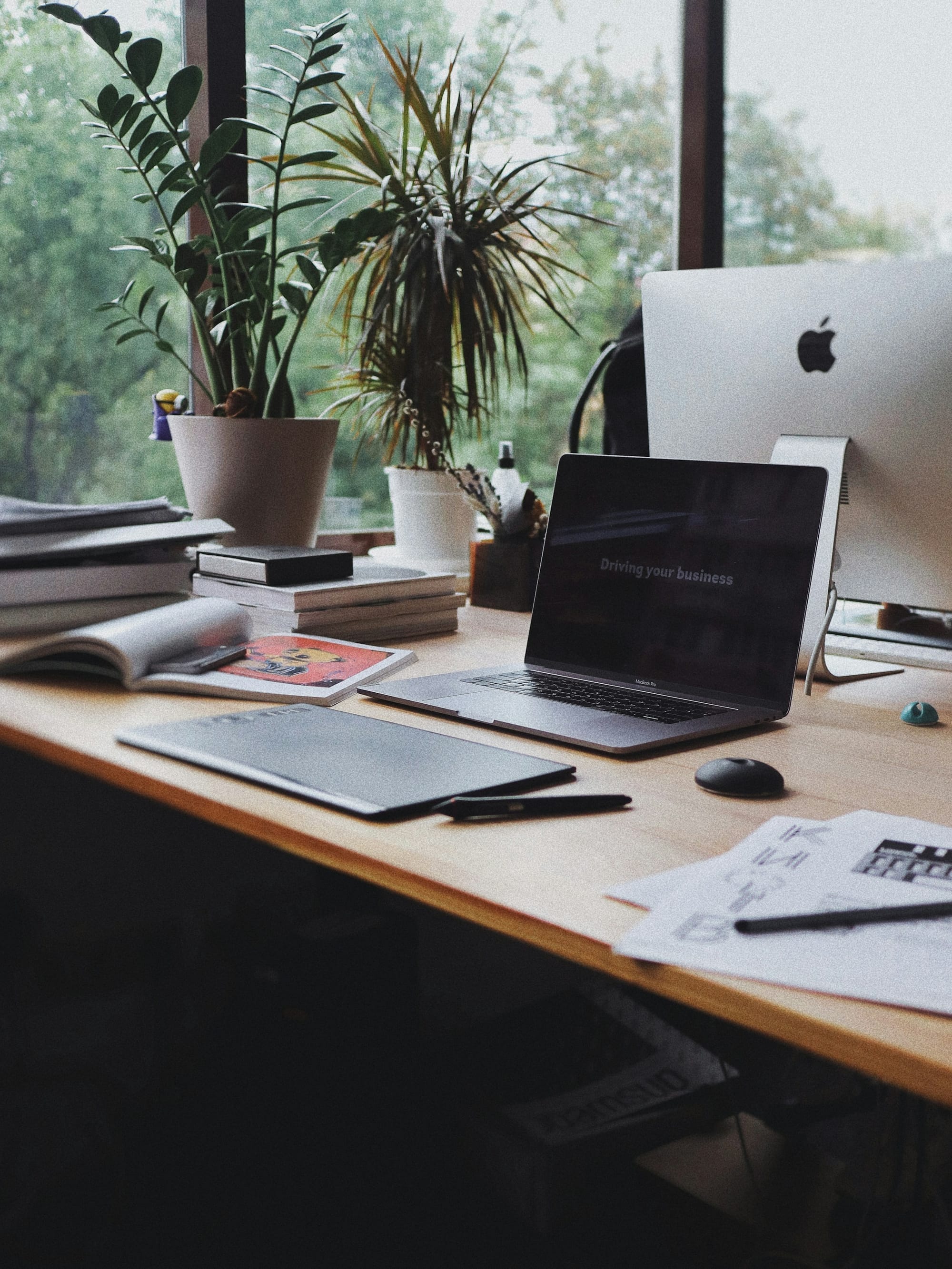 A plant-filled design studio desk with a laptop, desktop computer and printed materials.