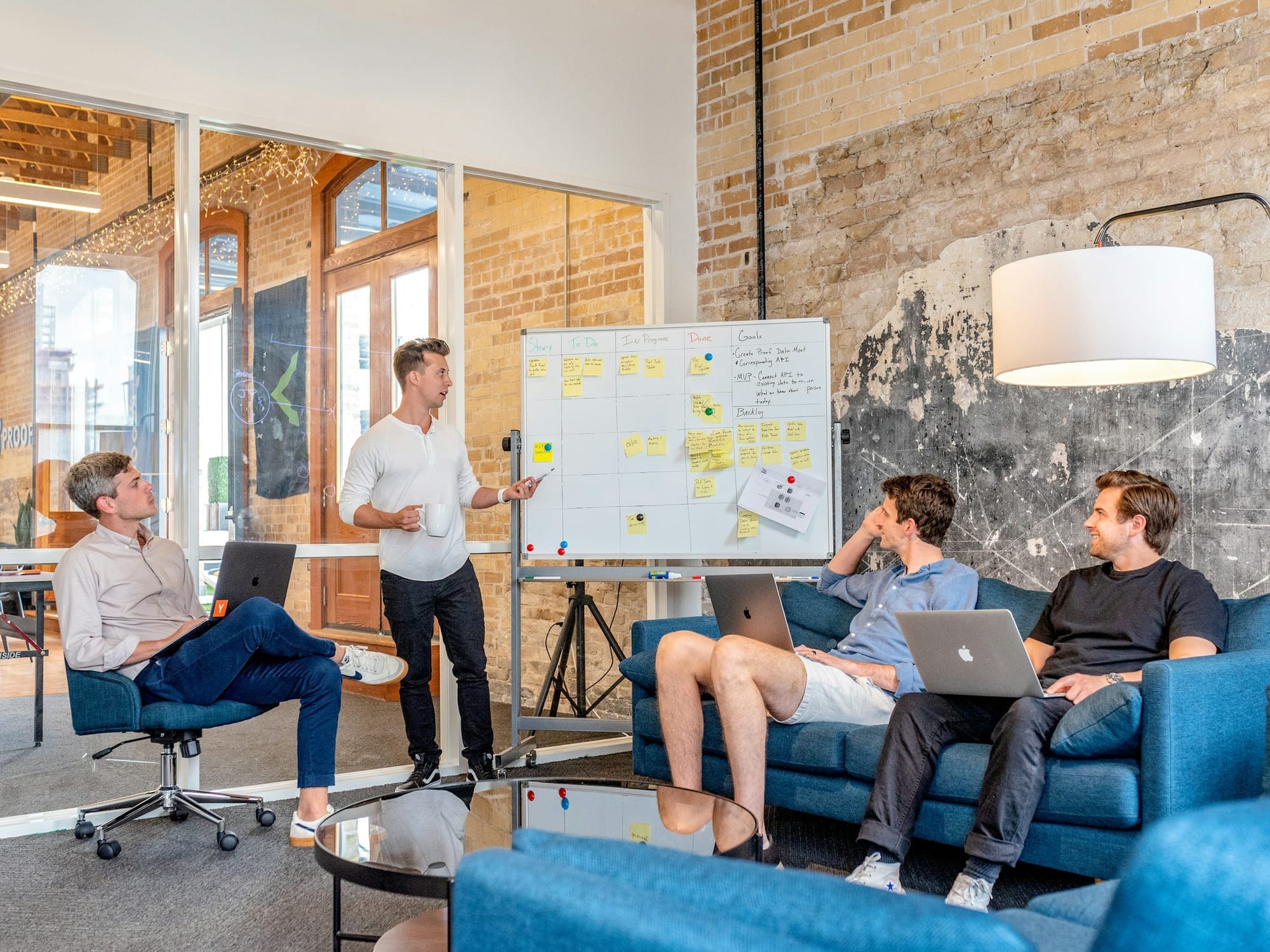 Four colleagues discussing work beside a whiteboard in a brick-walled office.