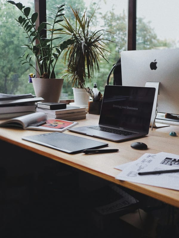 A plant-filled design studio desk with a laptop, desktop computer and printed materials.