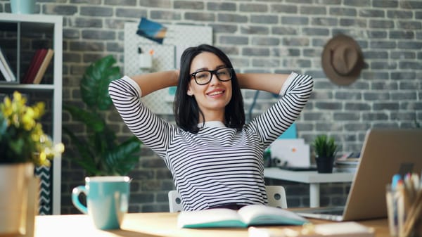 A smiling woman leaning back at her desk with her hands behind her head.